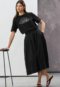 Tricou negru din bumbac cu imprimeu "Cannes", asortat cu o fustă neagră fluidă și sandale negre de plajă. În general, un look monocrom cu croi lejer.