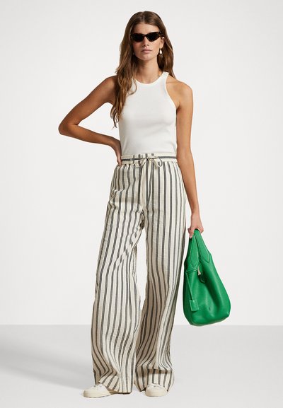 Débardeur blanc, pantalon large rayé en crème et noir, tenant un sac en cuir vert, associé à des lunettes de soleil noires.