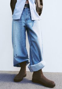 Jeans largi, decolorati, de culoare albastru, cu manșete răsucite, care prezintă ușoare uzuri. Asortați cu bocanci din suede maro închis, pe o suprafață gri.