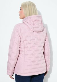 En blond kvinna med rakt hår bär en ljusrosa quiltad jacka med huva, sedd bakifrån mot en vit bakgrund.