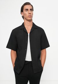 Black short-sleeve shirt with a Cuban collar, featuring a textured fabric and front button closure, paired with a white undershirt.