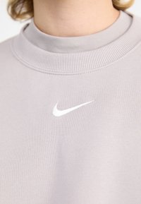 Grå hettegenser med ribbet rund hals og hvit brodert Nike swoosh-logo på venstre side. Myk stofftekstur.