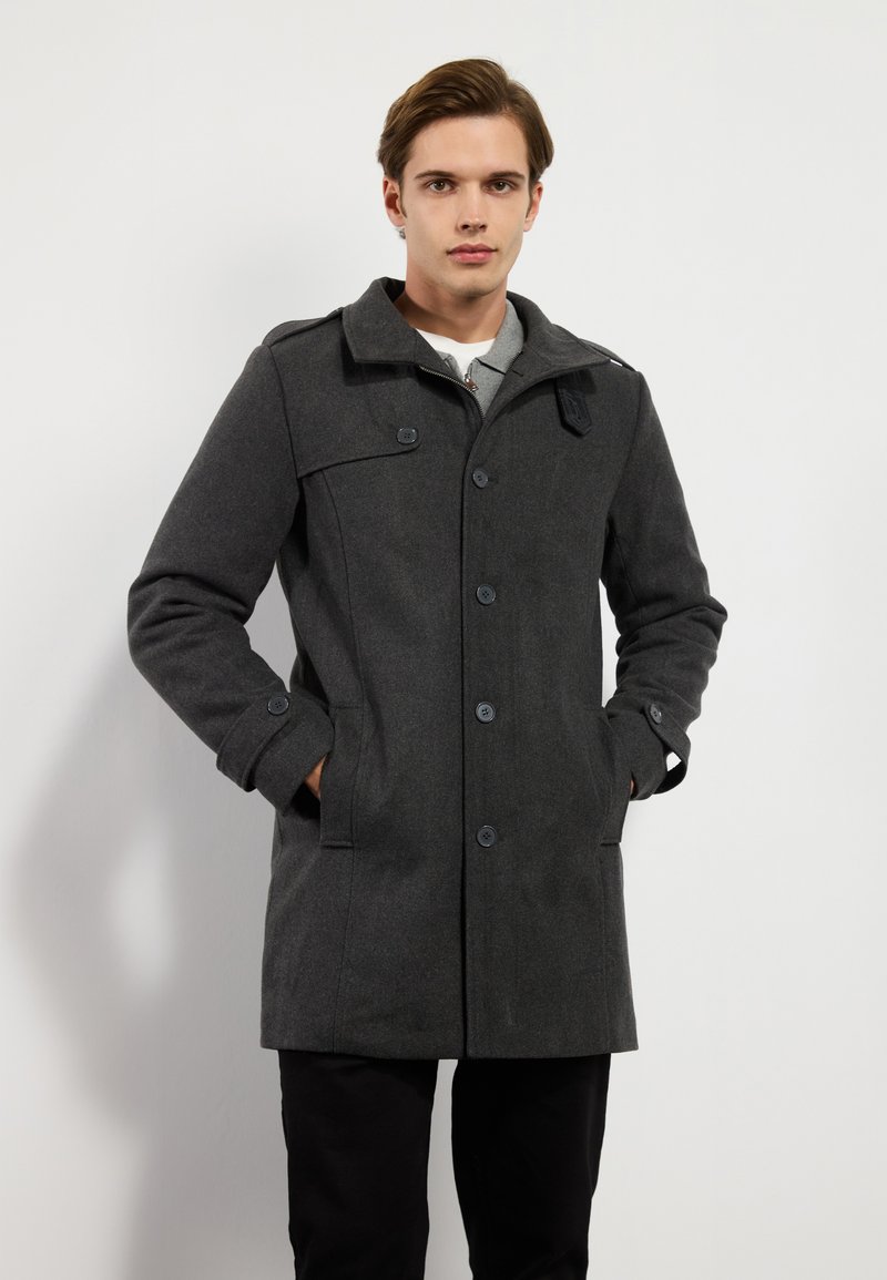 Cappotto grigio di lana con colletto alto, chiusura con bottoni, tasche frontali e dettagli sui gomiti, realizzato in un tessuto testurizzato e con vestibilità su misura.