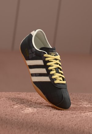 Sort Adidas sneaker med hvide striber, gule snørebånd og brun sål hvilende på tekstureret brun overflade.