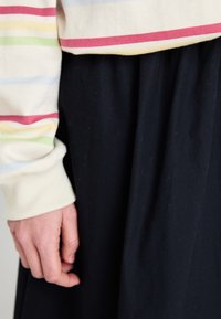 Stripete genser med lys kremfarge som har rosa, grønn og blå striper, kombinert med et flytende svart skjørt. Hånd hviler på skjørtet.