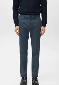 Man wearing dark blue slim-fit trousers, black loafers, and a black long-sleeve top, standing with arms relaxed at sides.