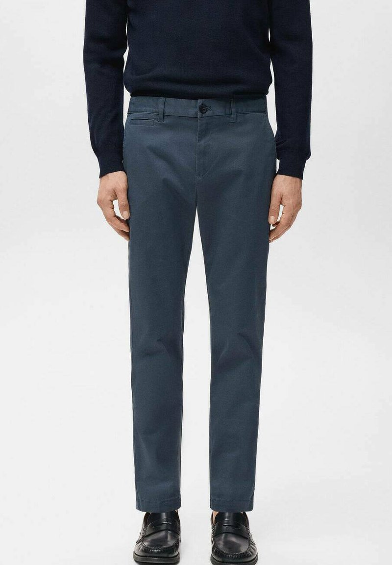 Man wearing dark blue slim-fit trousers, black loafers, and a black long-sleeve top, standing with arms relaxed at sides.