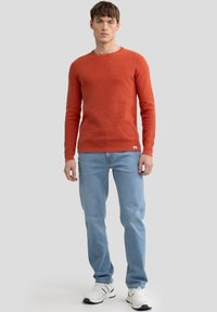 Rostfarbener, langärmliger, strukturierten Pullover, kombiniert mit hellblauen Jeans und weißen Sneakers, mit einem Rundhalsausschnitt und subtilen Branding-Details.