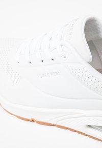 Weißer Sportschuh mit einer glatten Oberseite, perforierten seitlichen Details und weißen Schnürsenkeln. Verfügt über eine beige Gummisohle und ein strukturiertes Innenfutter.