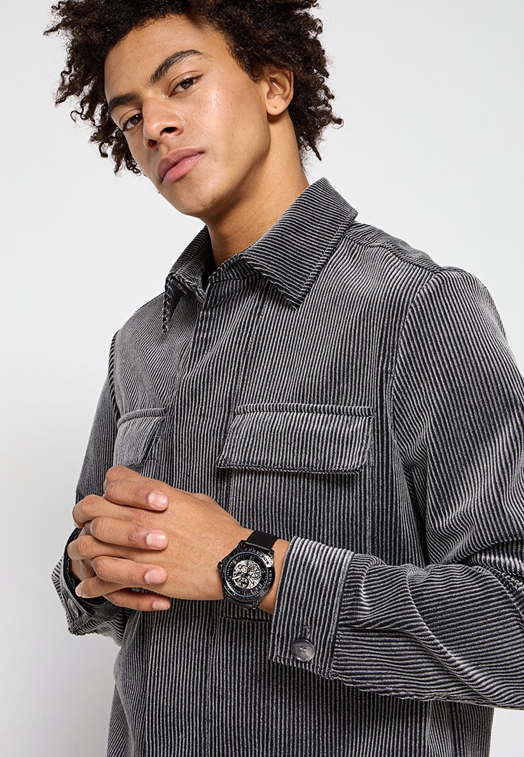 Camisa de pana a rayas blancas y negras con dos bolsillos en el pecho, puños abotonados y un reloj negro con un dial detallado.