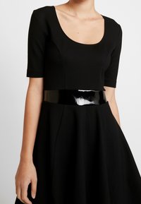 Svart klänning med korta ärmar, rundad halsringning, åtsittande liv och utsvängd kjol. Har ett glansigt svart lackbälte i midjan.