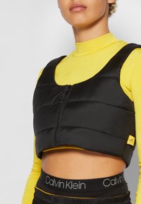 Personne portant un gilet court rembourré noir à fermeture éclair sur un haut jaune à col roulé, avec la ceinture noire Calvin Klein visible à la taille.
