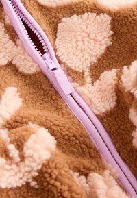 Fleece stof in bruin met grote lichtroze bloemenpatronen; voorzien van een roze ritssluiting met gedetailleerd textuur langs de rand.