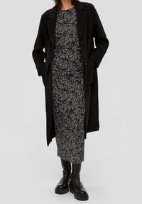 Schwarzer Mantel mit maßgeschneidertem Design, über einem knielangen Kleid mit einem weißen, abstrakten Muster getragen. Schwarze Stiefeletten mit dicken Sohlen.