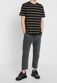 Camiseta de rayas azul marino con rayas amarillas, de corte holgado, combinada con jeans grises cortos y zapatillas negras. Atuendo simple y casual.