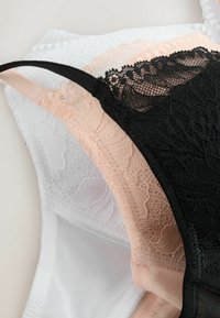 Pile de trois soutiens-gorge en dentelle noire, beige et blanche avec des motifs floraux et des bretelles réglables sur une surface blanche.