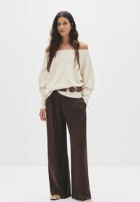 Cremefarbener schulterfreier Pullover, braune weite Hose mit Taschen, silberner runder Gürtelakzent, flache Schuhe mit Blumenmuster. Neutrale Hintergrund.