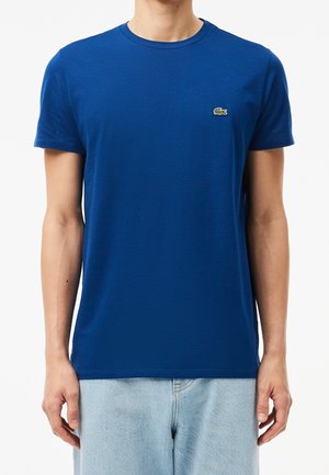 Homme portant un t-shirt bleu uni à manches courtes avec un petit logo crocodile vert sur la poitrine gauche et un jean bleu clair sur fond blanc.