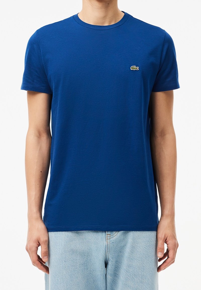 Homme portant un t-shirt bleu uni à manches courtes avec un petit logo crocodile vert sur la poitrine gauche et un jean bleu clair sur fond blanc.