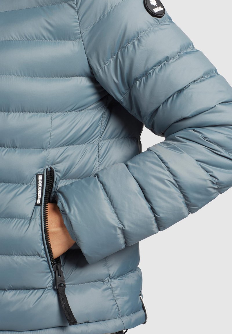 Helleblaue Daunenjacke mit gestepptem Design, ausgestattet mit einer Reißverschlusstasche und einem schwarzen Logo-Patch am Ärmel.
