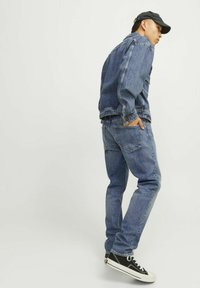 Jachetă din denim și blugi de nuanță albastru mediu, având o croială lejeră, textură ușor uzată și buzunare frontale. Adidași negri cu talpă albă.