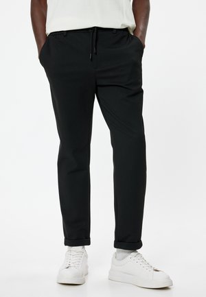 Sorte joggerbukser med elastisk talje og snor. Lavet af glat stof, med sidelommer og opsmøgte bukseben. Brugt med hvide sneakers.