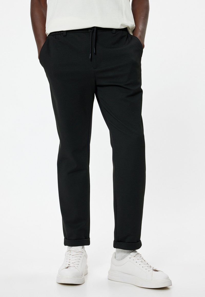Jogging noir avec une taille élastique et un cordon de serrage. Fabriqué en tissu lisse, avec des poches latérales et des ourlets retroussés. Porté avec des baskets blanches.