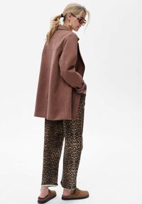 Bruine oversized jas, panterprint broeken en beige instapschoenen. De jas heeft een gladde textuur met een kraag en zakken.