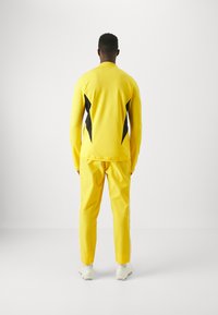 Chándal deportivo amarillo con acentos laterales negros. Parte superior de manga larga y pantalones ajustados. Tejido suave y rayas en contraste en las piernas.