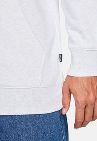 Sudadera blanca con tejido texturizado, puños y dobladillo acanalados. Presenta un bolsillo lateral y una pequeña etiqueta negra de la marca en la parte inferior.