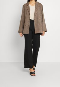 Hnědý kostkovaný dvouřadový blejzer, oversized střih, v kombinaci s přiléhavým béžovým topem a černými kalhotami s vysokým pasem a širokými nohavicemi. Černé sklouzávací sandály.