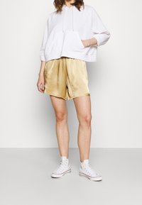 Weißer Kapuzenpullover mit Kängurutasche, kombiniert mit glänzenden goldenen Shorts. Das Model trägt weiße High-Top-Sneaker. Minimalistischer Hintergrund.