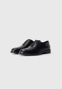 Une paire de chaussures oxford en cuir verni noir avec un laçage fermé et des talons bas sur fond blanc.