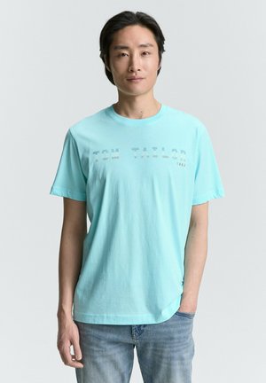 Camiseta de algodón azul claro con mangas cortas que presenta "TOM TAYLOR 1982" impreso en un diseño sutil y contrastante. Corte casual.