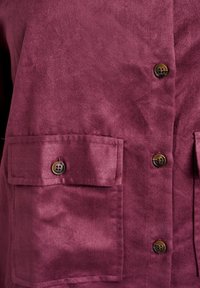 Burgundowa koszula o zamszowej fakturze, z dwiema przednimi kieszeniami z zapięciem na guziki oraz rzędem guzików pośrodku do zapinania.