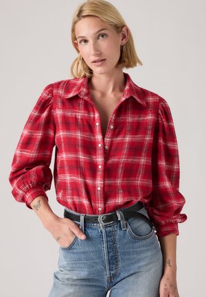 Femme aux cheveux blonds courts portant une chemise à carreaux rouge boutonnée et un jean bleu clair avec une ceinture noire sur un fond uni.