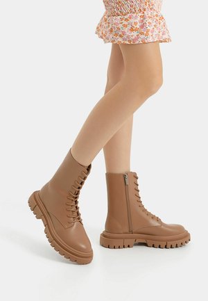 Piernas vestidas con una falda floral y botas marrones con cordones gruesos y cremallera lateral, de pie sobre un fondo blanco.
