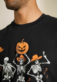 Sort bomulds T-shirt med farverige skeletter med hatte og en orange græskar grafik. Blød tekstur med korte ærmer.