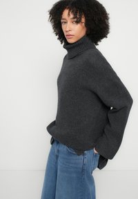 Pulover cu guler înalt tricotat, de culoare gri închis, cu croială lejeră, guler răsucit și tăieturi laterale, combinat cu blugi evazați de culoare albastru deschis.