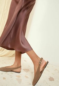 Scarpe slingback in suede marrone con punta appuntita, caratterizzate da un accento a cerchio in metallo e una suola piatta, abbinate a una gonna in satin marrone.