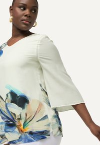 Cremefarvet bluse med blomsterprint, der har V-udskæring og klokkeærmer. Stoffet er glat og prydet med blå og beige blomstermønstre på fronten.