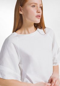 Joven con cabello largo y rojo lleva una camiseta blanca de manga corta y lisa, mirando hacia un lado sobre un fondo blanco.