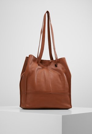 Sac tote en cuir marron avec longues bretelles doubles, texture douce et base rectangulaire structurée sur un piédestal blanc.