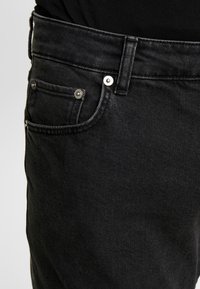 Jeans de mezclilla negra con una vista en primer plano de la cintura y el área del bolsillo. Presentan herrajes plateados, una textura sutil y un diseño clásico de cinco bolsillos.