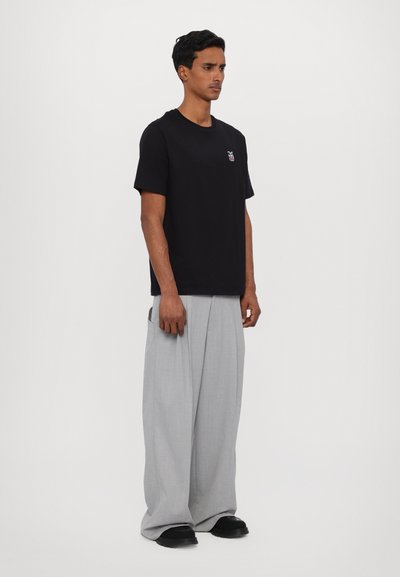 T-shirt en coton noir à manches courtes avec un petit logo coloré sur la poitrine gauche ; associé à un pantalon gris ample à plissage.