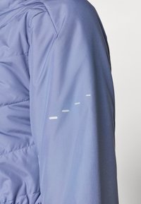 Jachetă puffer violet deschis cu textură matlasată. Dispune de dungi reflective pe mânecă. Material neted și siluetă ajustată.