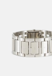 Montre en acier inoxydable argenté avec un boîtier rectangulaire et un bracelet à maillons. Présente une gravure "GUESS" et une finition polie.