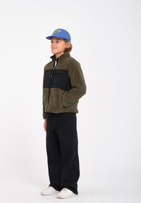Veste en polaire verte avec panneau avant noir, fermeture éclair, deux poches avant ; associée à un pantalon large noir et des baskets blanches. Casquette bleue sur la tête.