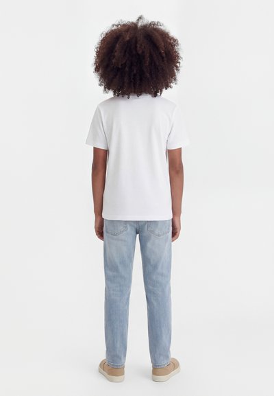 Copil cu păr creț purtând un tricou alb simplu, blugi albastru deschis și pantofi bej, stând cu spatele la un fundal alb.
