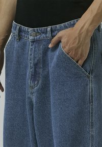 Blue denim jeans featuring a high waist, loose fit, visible stitching, and a front button closure, with hands in side pockets.